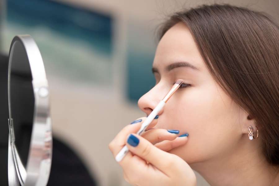 Une jeune femme applique délicatement du fard à paupières à l'aide d'un pinceau fin en se regardant dans un miroir pour fixer son maquillage.