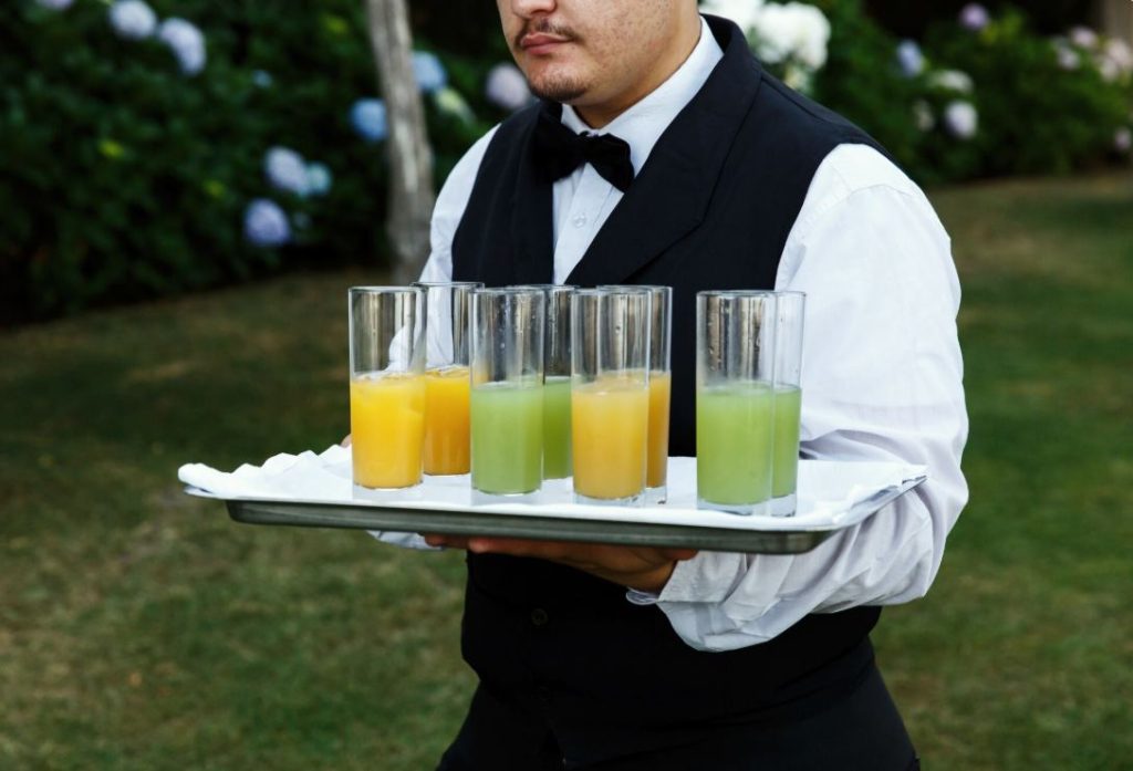 Serveur en tenue élégante portant un plateau de verres colorés illustrant des idées mocktails et boissons sans alcool mariage.